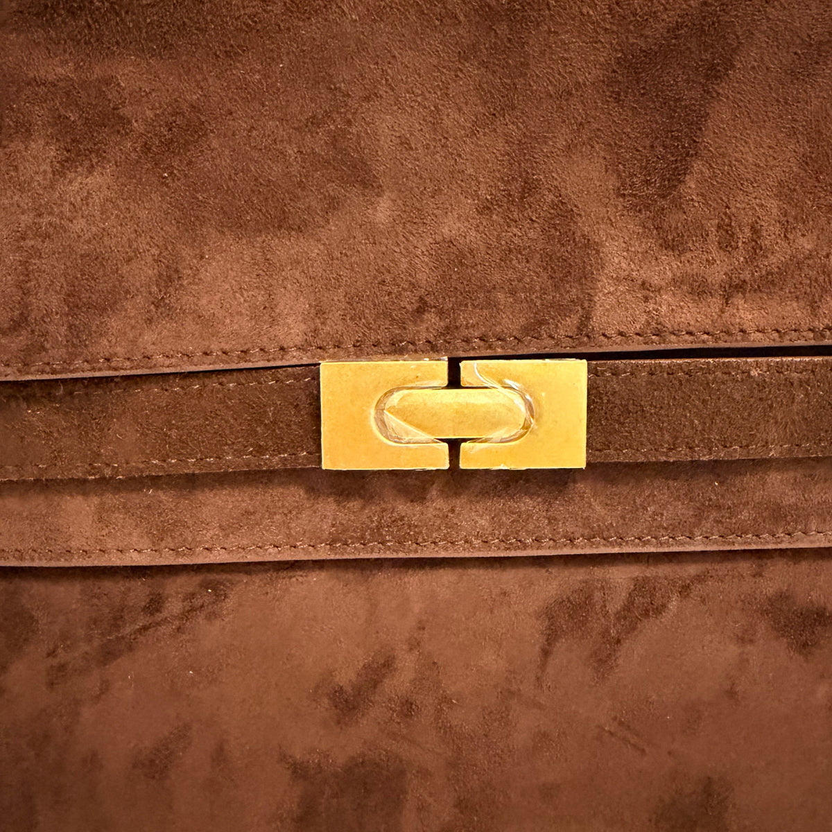 Close-up of a brown suede or nubuck bag featuring a vertical strap with a polished gold-tone metal clasp.