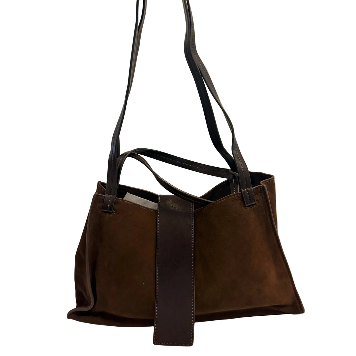A dark brown suede tote bag with black leather handles and a vertical black leather strap detail, displayed on a white surface against a light gray wall.