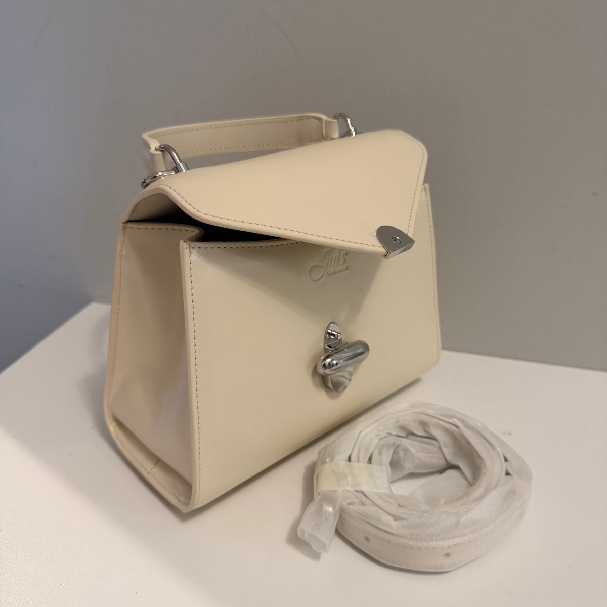 An off-white leather handbag with silver-tone hardware and a matching detached shoulder strap, displayed on a white surface against a gray wall.