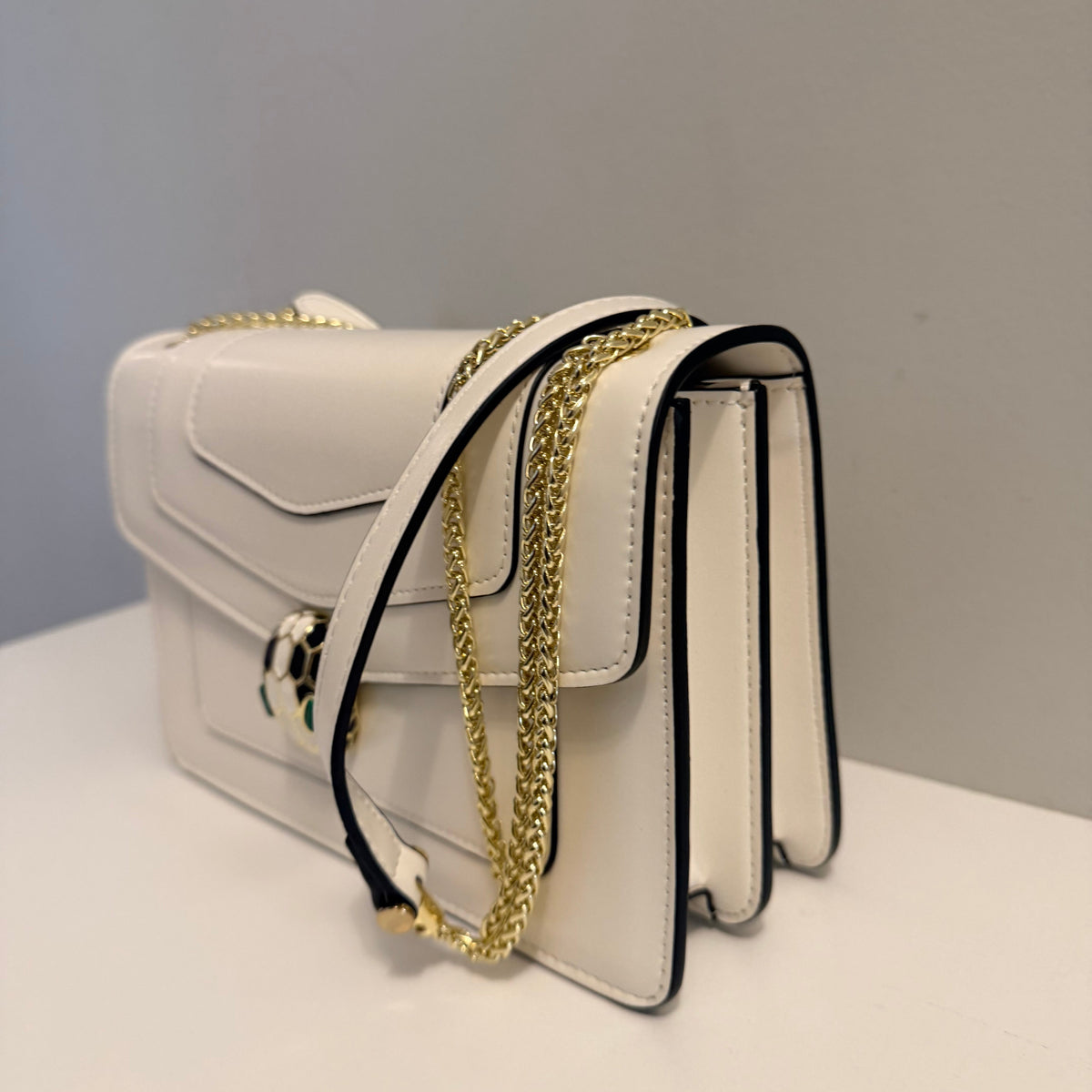 A white faux-leather shoulder bag with black piping details and a gold chain strap, displayed on a white surface against a gray background.
