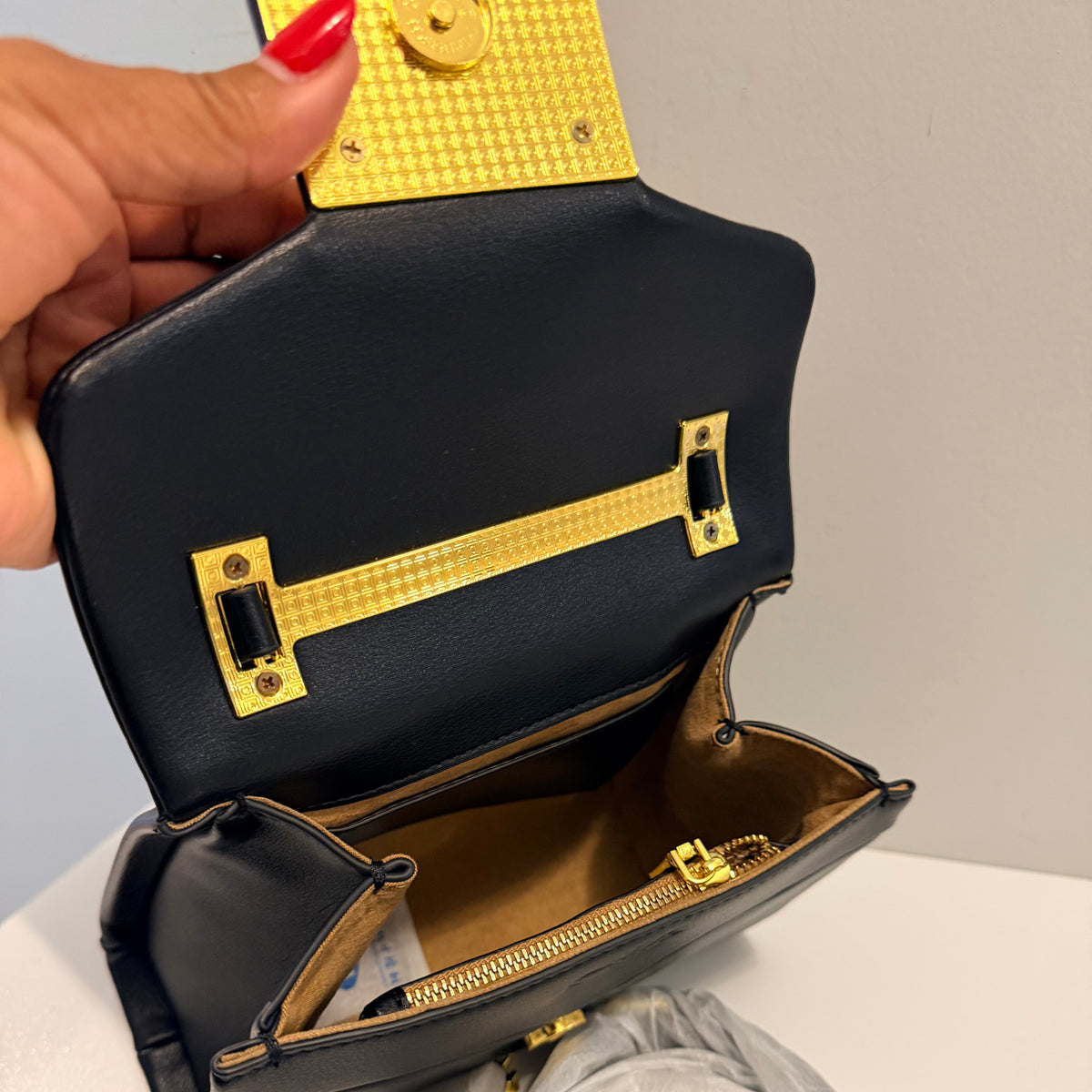 A black leather handbag with gold-tone hardware is shown open, held by a hand with red nail polish. A light gray fabric dust bag is visible next to it on a white surface.