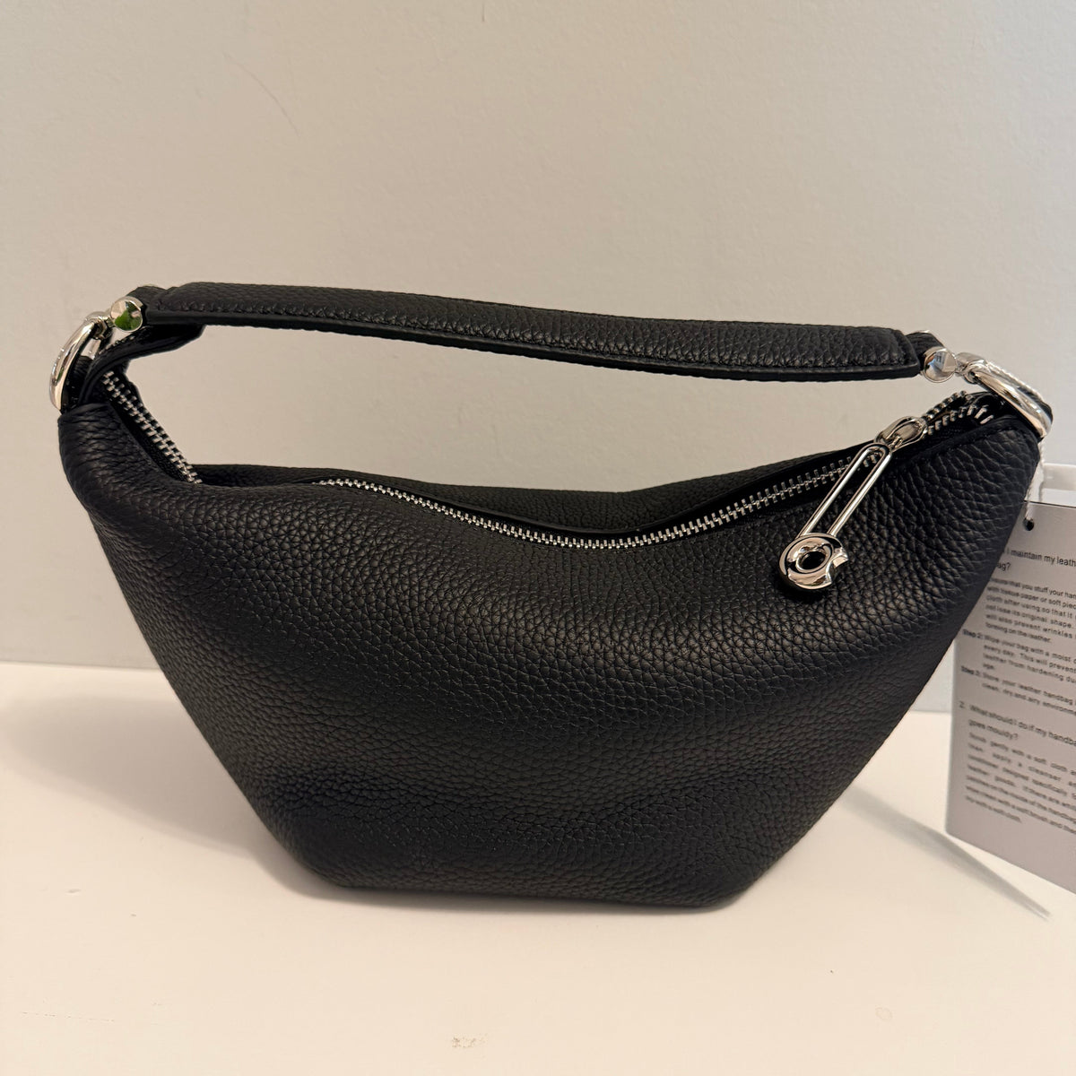 A black pebbled leather shoulder bag with silver-tone hardware, shown against a plain off-white background with a paper tag attached.