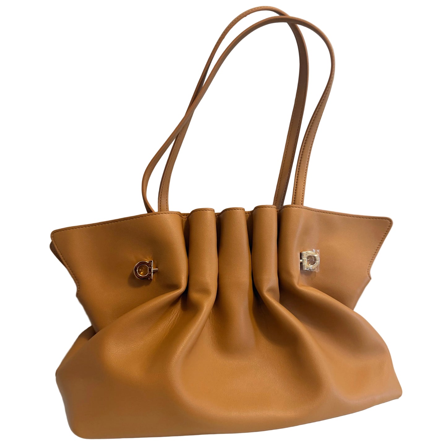 A tan leather handbag with a gathered, slouchy silhouette, dual shoulder straps, and small gold-tone hardware, displayed on a white surface against a light gray wall.
