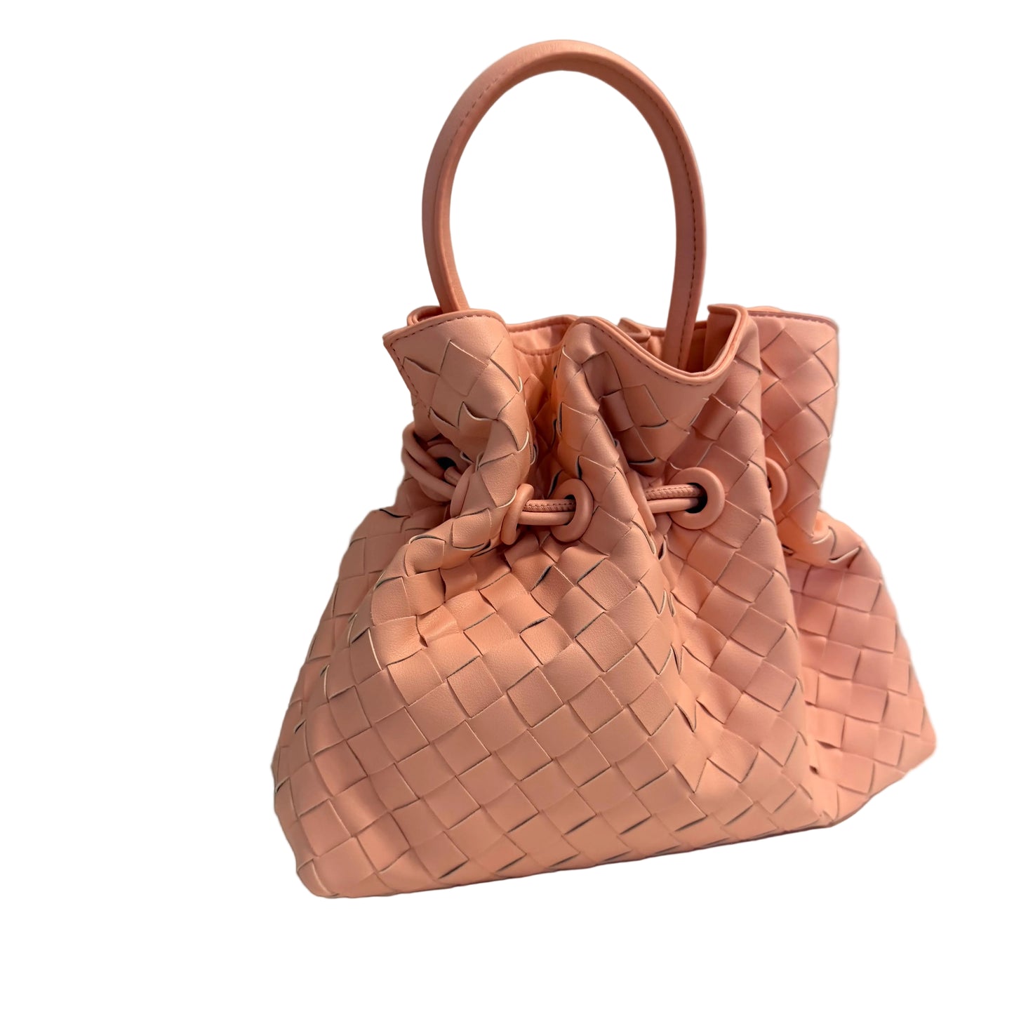 A light pink woven leather bucket bag with a single top handle, resting on a white surface against a light gray wall.