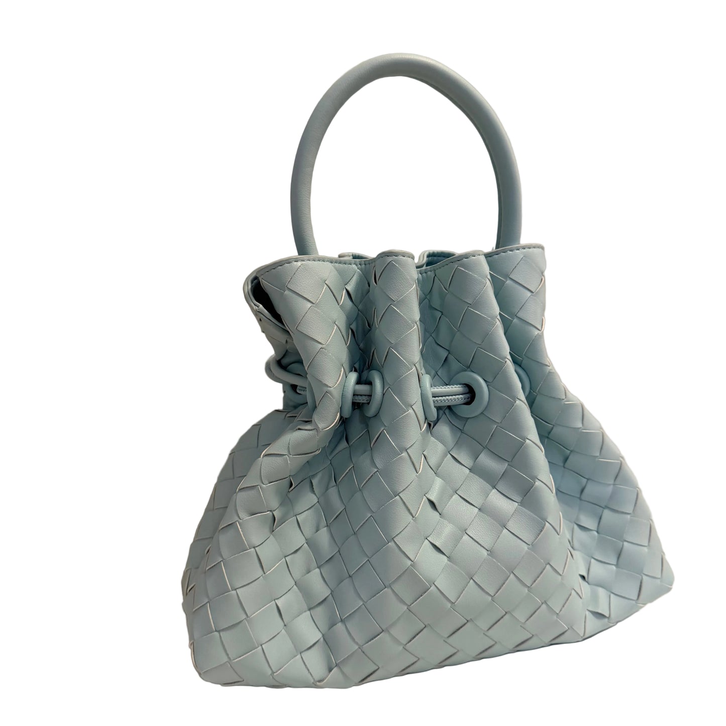 A light blue woven leather bucket bag with a single top handle, resting on a white surface against a plain light gray wall.