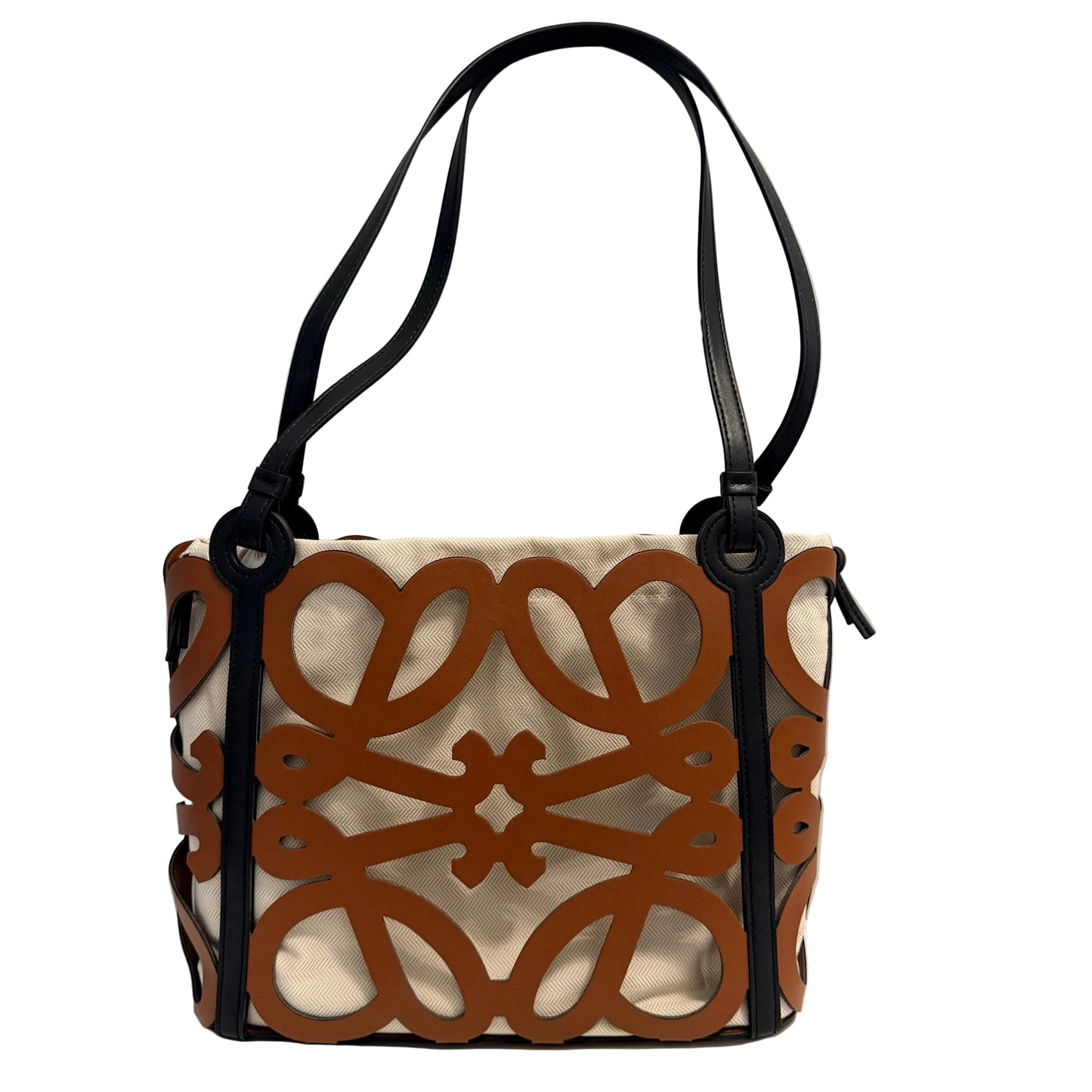 A structured tote bag with a cream canvas body, brown interlocking geometric cutout pattern on the front, and black leather straps and trim, shown on a white surface against a light gray wall.