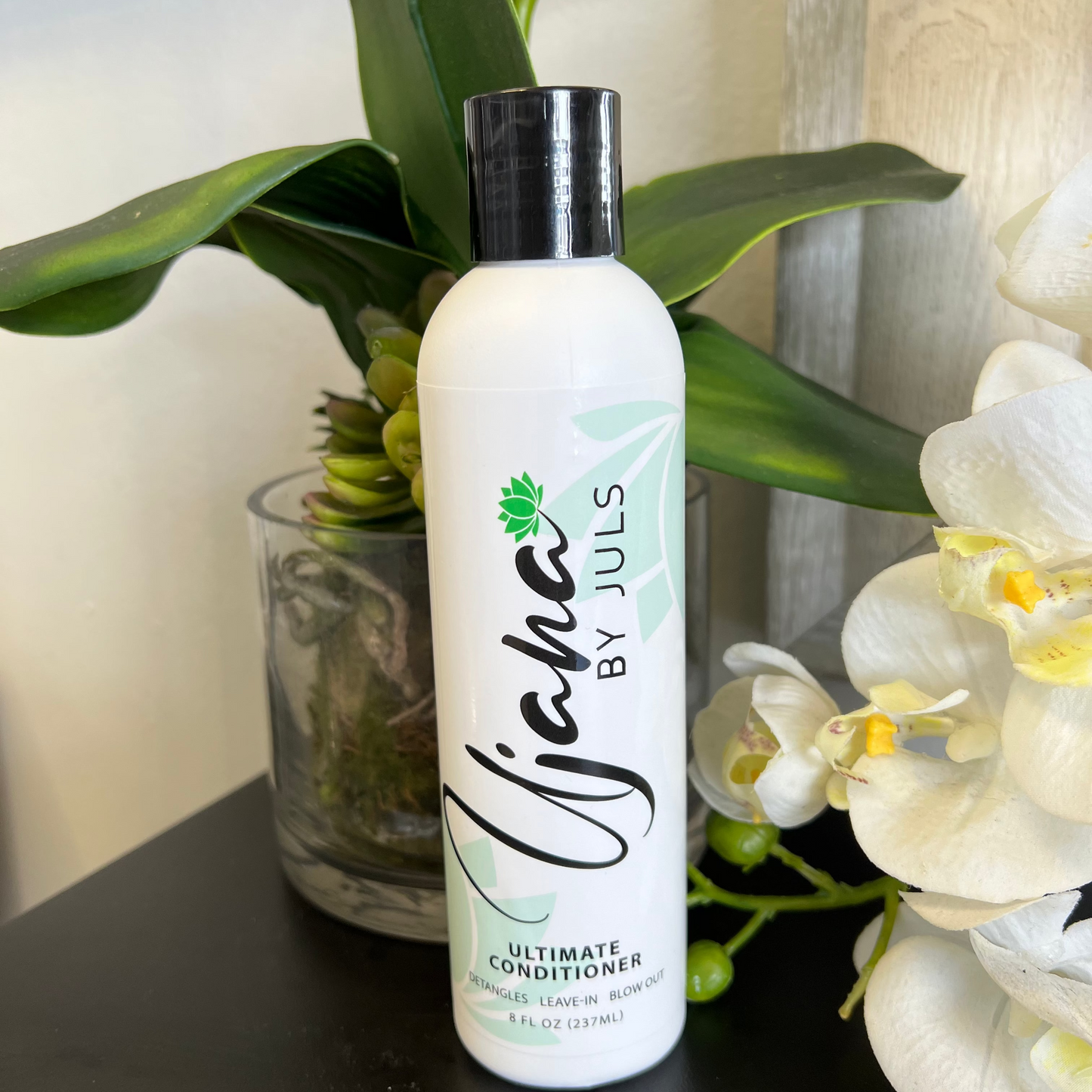 A bottle of Ujana Ultimate Conditioner with a white and green label, displayed on a table with a plant in the background.
