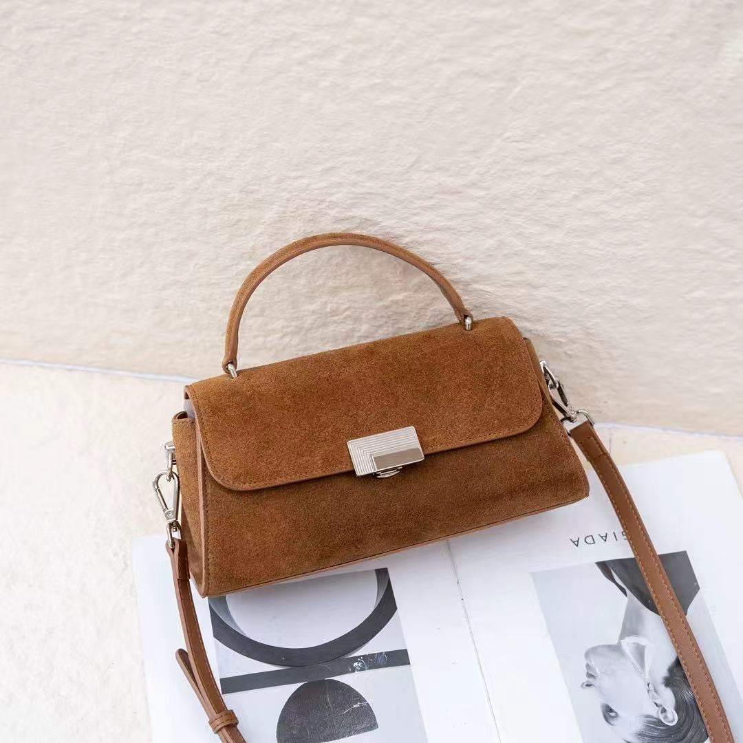 A brown suede handbag with a top handle and a silver-tone clasp, resting on an open magazine against a textured off-white wall.