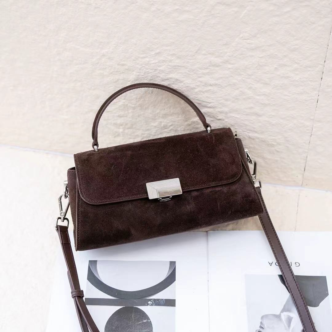 A dark brown suede handbag with a top handle, a silver-tone rectangular clasp, and a detachable shoulder strap, styled on top of open magazine pages against a textured off-white background.