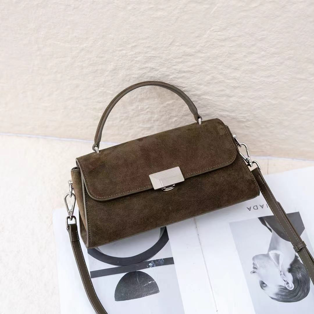 A dark brown suede handbag with a top handle and a silver-tone clasp, resting on a magazine page against a textured off-white wall.