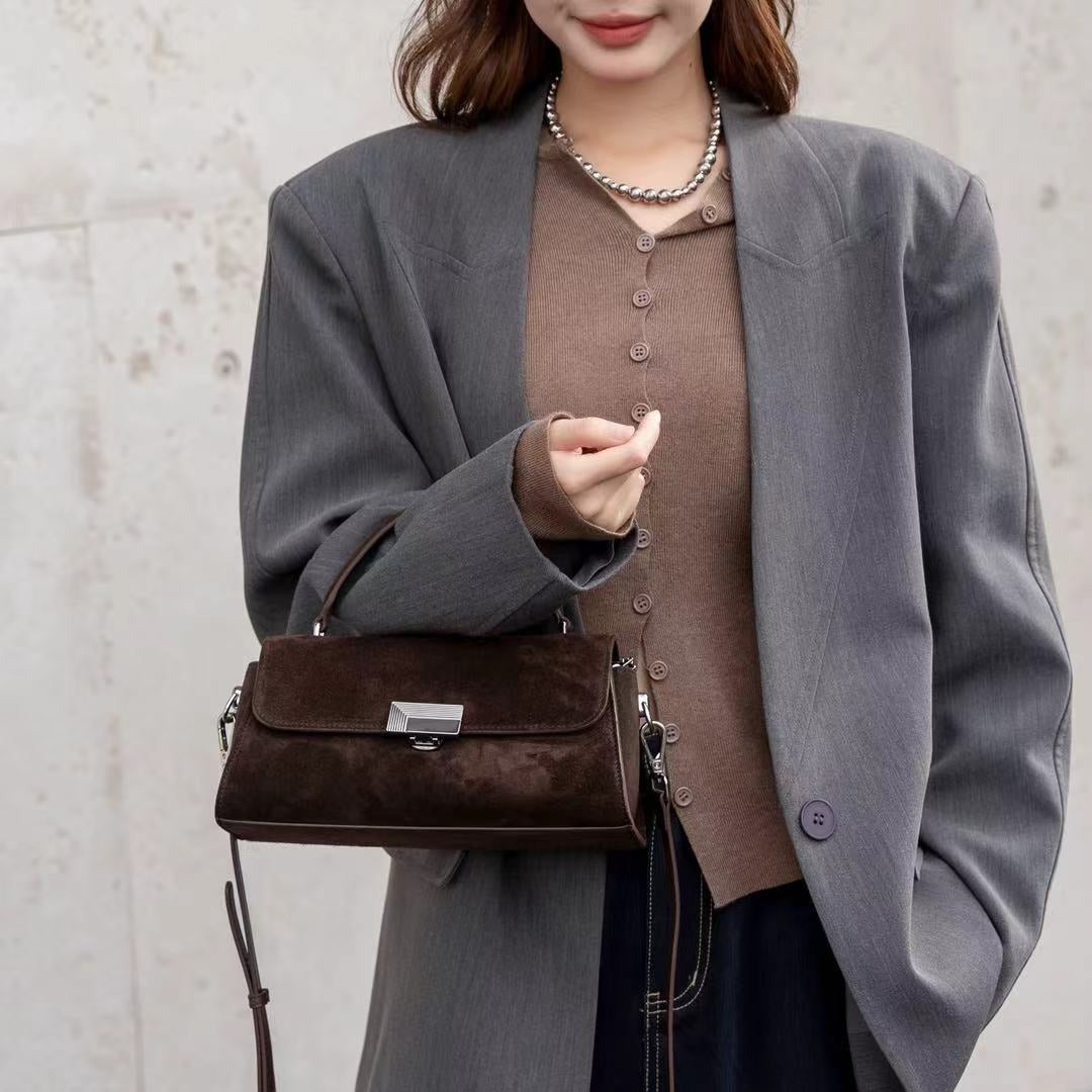 A woman wearing an oversized gray blazer over a brown button-front top and dark wide-leg pants, carrying a structured dark brown handbag with a long strap against a light concrete wall.