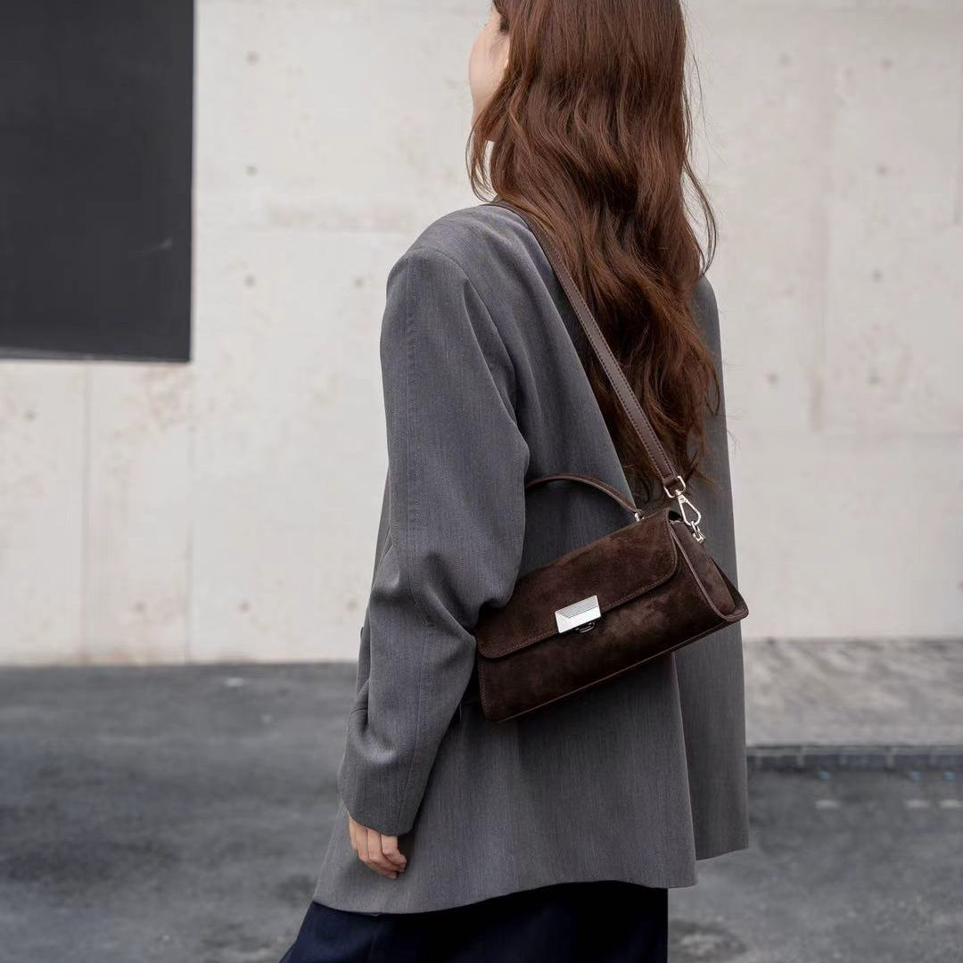 A woman with long brown hair is shown from behind wearing a dark gray blazer and dark navy wide-leg pants, carrying a dark brown textured leather shoulder bag with a silver-toned clasp.