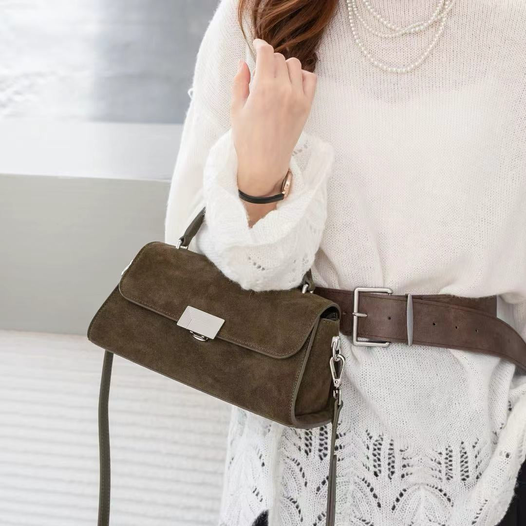 A person wearing a white knit sweater and black skirt carries a dark brown suede shoulder bag with a long strap and silver-tone hardware.