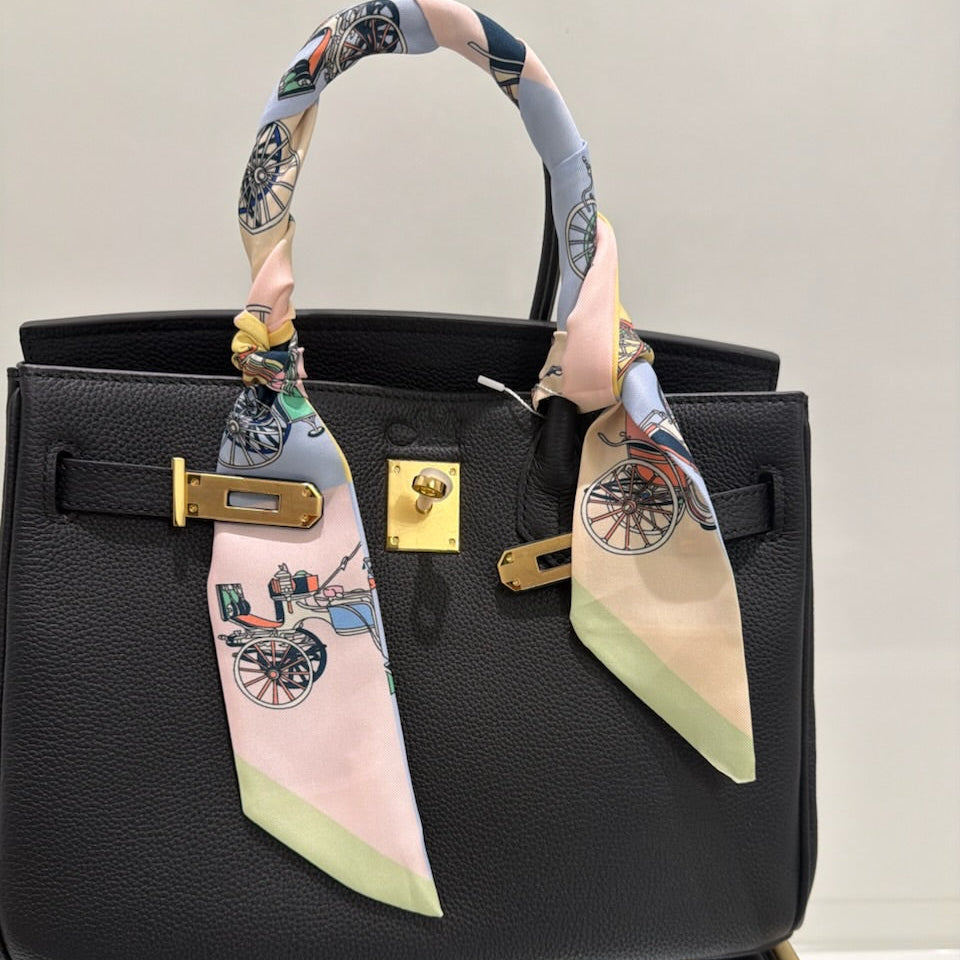 Black handbag with a colorful scarf draped over the handle on a reflective surface.