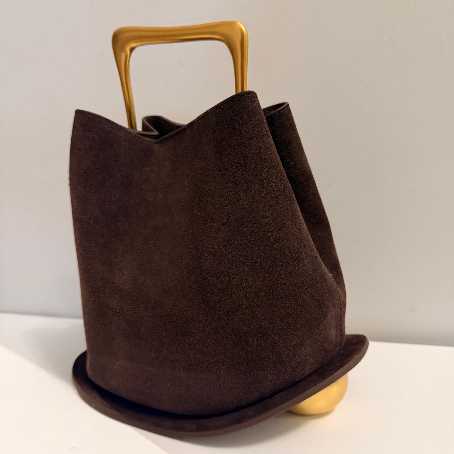 Brown suede handbag with gold handle and base on a light background