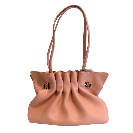 A light pink leather shoulder bag with gathered pleats and double top handles, featuring gold-tone metal hardware, set against a plain light gray wall and white surface.