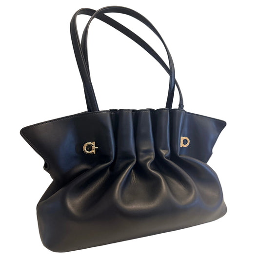 A black leather slouchy handbag with double shoulder straps and gold-tone metal hardware, displayed on a white surface against a light gray wall.