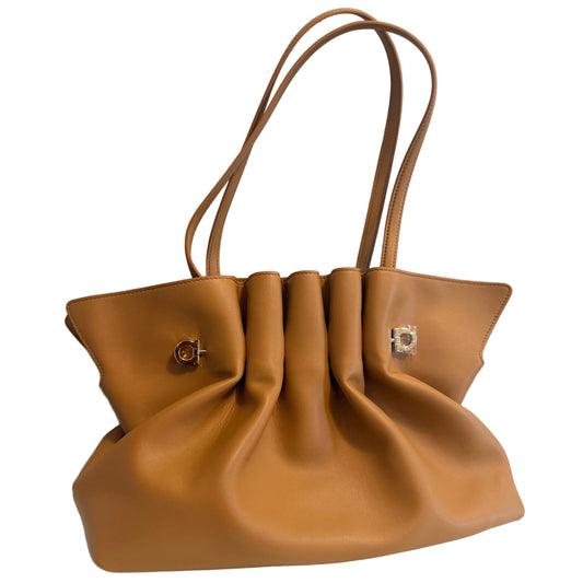A tan leather handbag with a gathered, slouchy silhouette, dual shoulder straps, and small gold-tone hardware, displayed on a white surface against a light gray wall.