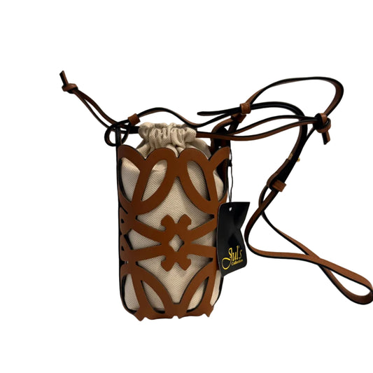 A brown leather crossbody bag with a geometric cutout pattern over a cream canvas pouch, featuring a drawstring top, a black hangtag with gold lettering, and a long brown strap, set against a plain light gray background.