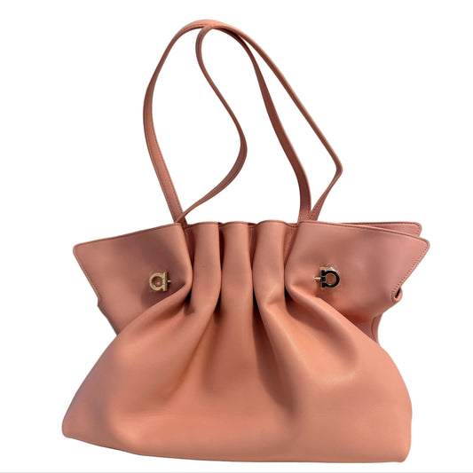 A dusty pink leather handbag with a gathered, slouchy silhouette, dual shoulder straps, and small gold-tone hardware, displayed on a white surface against a light gray wall.