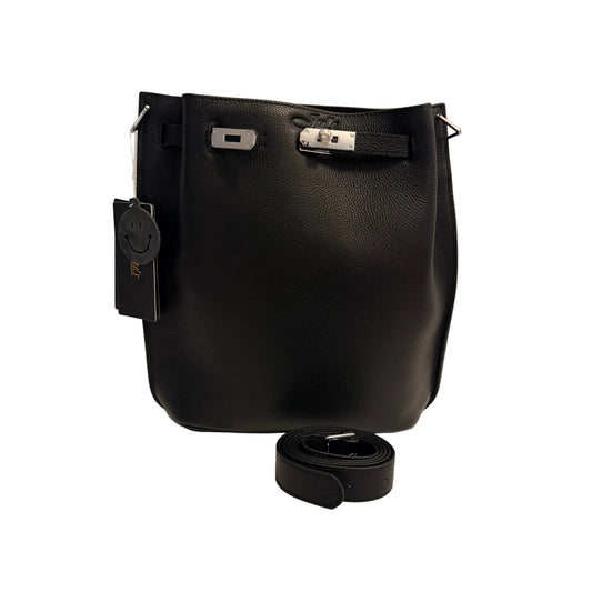 A black pebbled leather bucket bag with silver-tone hardware, shown on a white surface against a light gray wall. A black hangtag is attached on the left, and a matching black leather strap is coiled in front of the bag.