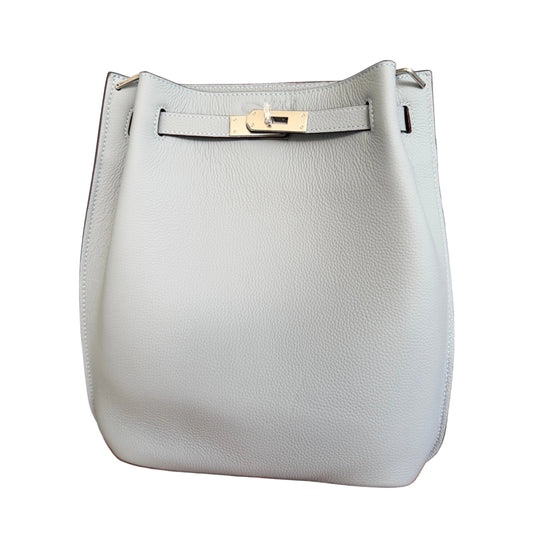A light blue pebbled leather bucket bag with gold-tone hardware, displayed on a wooden surface against a gray wood-paneled wall.